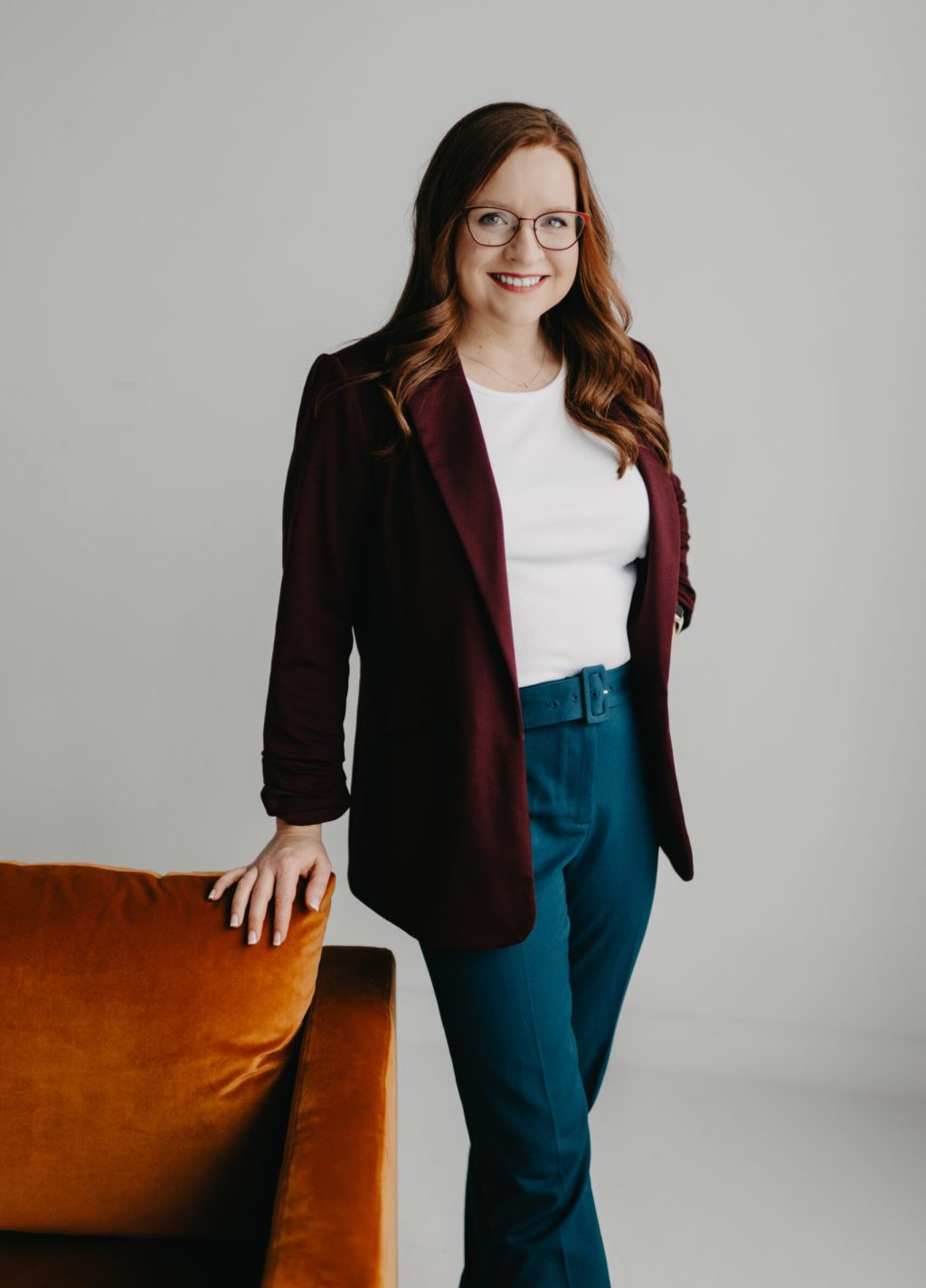 jordyne carmack, full body image, woman wearing maroon blazer and a white shirt, confident with one hand on hip. orange chair.